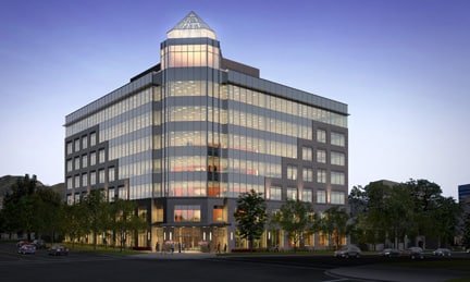 A well lit glass business building with a tower in the front has cars parked outside at twilight